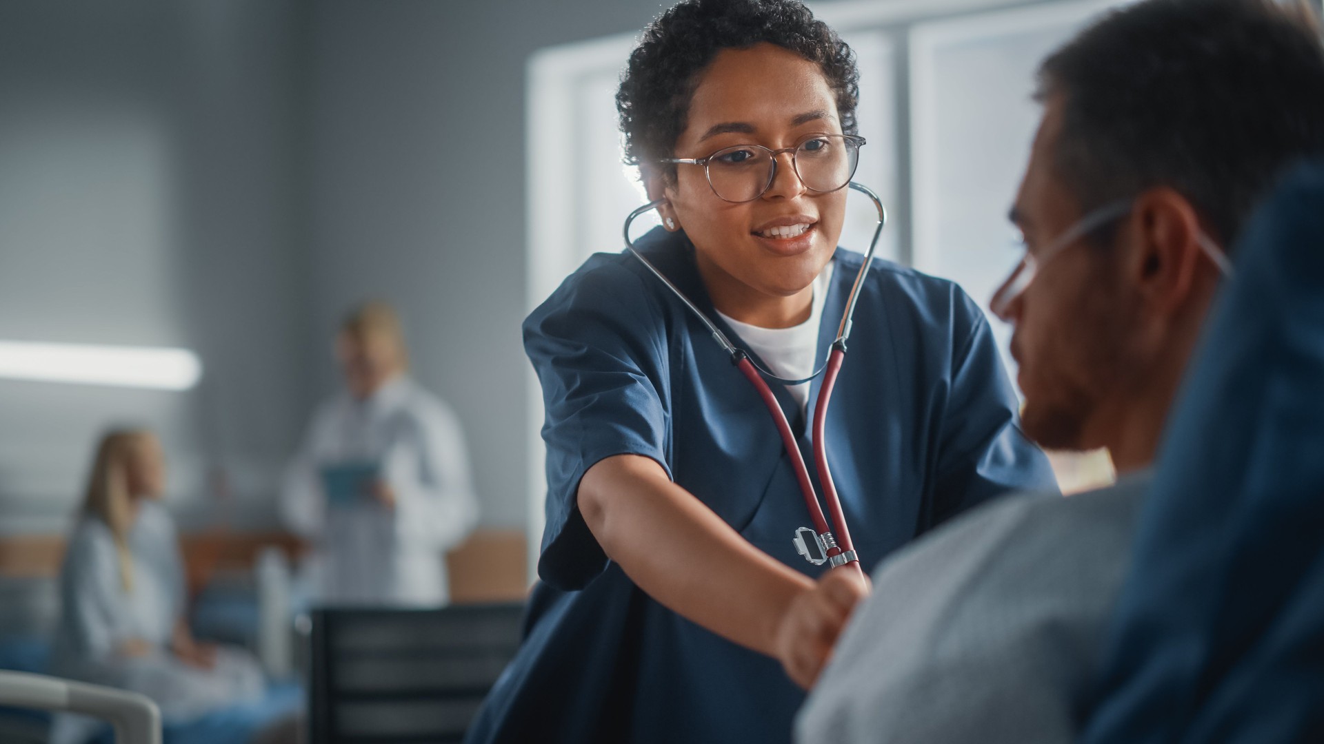 Hospital Ward: Friendly Black Head Nurse Uses Stethoscope to Listen to Heartbeat and Lungs of Recovering Male Patient Resting in Bed, Does Checkup. Man Getting well after Successful Surgery